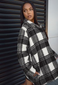 Överdimensionerad fleecejacka i svart och vitt rutmönster med krage, med en mjuk struktur och sidofickor. Ärmarna är något vikta.