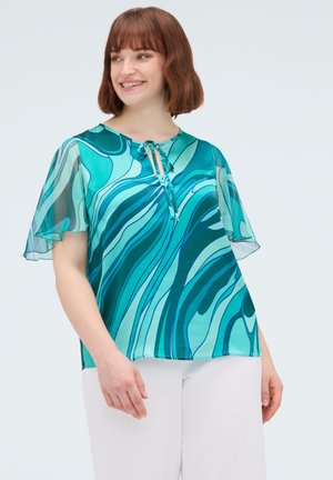 Vrouw glimlacht met een blouse met teal- en blauwe spiraalprint, doorschijnende wapperende mouwen en een witte broek tegen een lichte achtergrond.