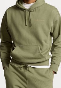 Olivově zelená mikina s kapucí vyrobená z měkké tkaniny, s přední kapsou typu kangaroo a malým vyšitým logem na hrudi.