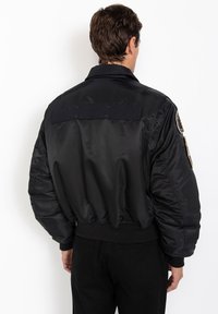 Giacca bomber nera con una texture liscia e lucida, polsini e vita a costine. Presenta una patch in velcro e dettagli con logo sulla manica.