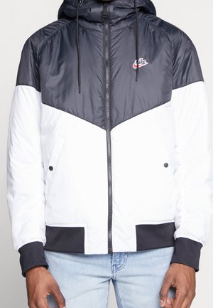 Homme portant une veste à capuche Nike noire et blanche avec fermeture éclair sur le devant et un jean bleu clair, visible du cou aux hanches.