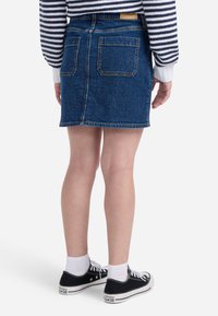 Denim mini krilo v temno modri barvi z vidnimi šivi, dvema zadnjima žepoma in ravnim krojem. Nosi se z črnimi Converse športnimi copati in belimi nogavicami.