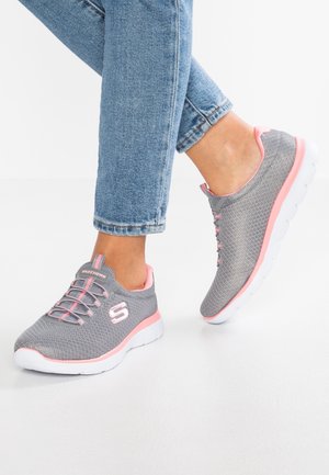 Legs with light blue denim jeans wearing gray and pink Skechers sneakers with white soles on a white surface.