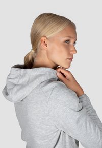 Felpa grigia chiara con cappuccio, caratterizzata da un tessuto texturizzato e un dettaglio con zip sul collo, mostrata di lato su uno sfondo neutro.