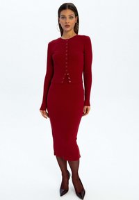 Czerwona sukienka prążkowana z długimi rękawami, zapięcie na guziki z przodu, dopasowany krój oraz długość do połowy łydki, zestawiona z szpilkami.