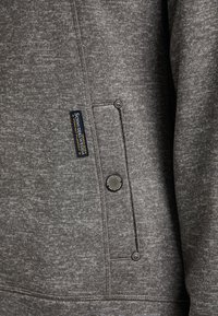 Sweatshirt gris en tissu texturé, avec une poche hip, des accents de boutons et une étiquette de marque cousue sur la couture latérale.