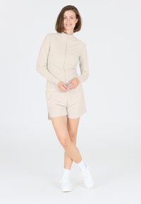 Helle beige Zip-Up-Jacke mit langen Ärmeln, kombiniert mit passender Shorts. Verfügt über eine glatte Textur und einen elastischen Taillenbund, ergänzt durch weiße Sneakers.