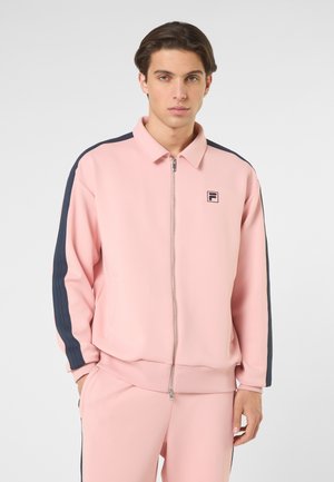 Joven con cabello oscuro que lleva un chándal rosa claro con cremallera y rayas oscuras en las mangas, junto con pantalones a juego, mirando hacia adelante.