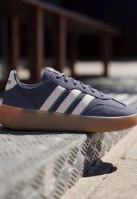 Zapatilla deportiva azul oscuro con rayas blancas y suela de goma descansando sobre un escalón metálico texturizado en un entorno exterior con fondo desenfocado.