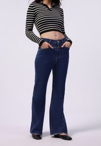 Jeans acampanados de color azul con cintura alta, que cuentan con un cierre de botón en la parte frontal y bolsillos. Combinados con una camiseta corta a rayas en blanco y negro.
