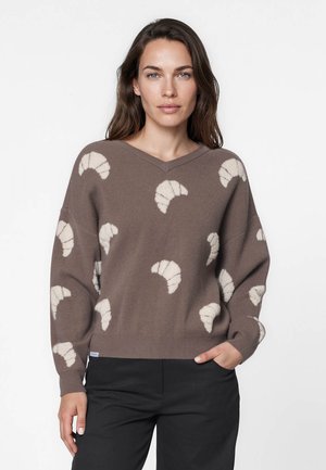 Femme aux longs cheveux portant un pull marron à motifs de croissants blancs, pantalon noir, debout avec une main dans la poche sur un fond uni.