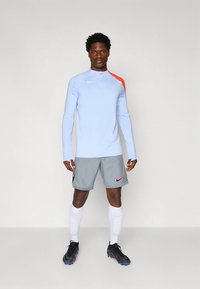 Vaaleansininen pitkähihaurheilupaita, jossa on punainen korostus, yhdistettynä harmaisiin shortseihin, joissa on pieni logo. Mustat lenkkarit ja valkoiset sukat.