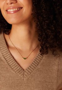 Gouden ketting met drie groene stenen accenten, gedragen om een blote nek, gecombineerd met een lichtbruine gestructureerde v-hals trui.