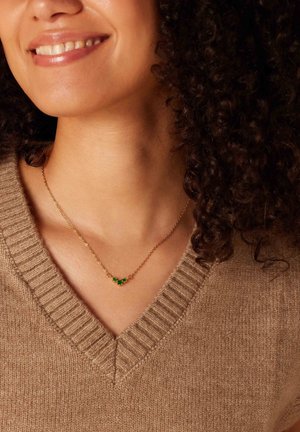Collier en chaîne dorée avec trois accents en pierre verte, porté sur un cou nu, associé à un pull en v-texturé beige clair.