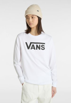 Young person wearing a beige knit beanie, white long-sleeve shirt with black Vans logo, and light-colored pants, standing against a plain background.