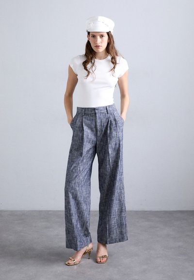 Weißes, geripptes, eng anliegendes Oberteil, kombiniert mit hochgeschnittenen, weiten Hosen aus strukturiertem blauen Stoff. Goldene, verstellbare Absätze runden den Look ab.