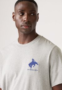 Grijze katoenen T-shirt met een ronde hals, voorzien van een blauw geborduurd logo van een cowboy en de tekst "Levi Strauss & Co." op de linkerkant van de borst.