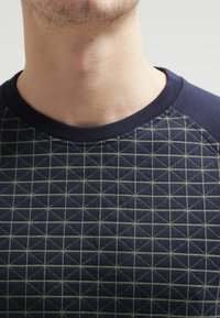 Primer plano del cuello y la parte superior del pecho de un hombre que lleva una camisa azul marino con un patrón de cuadrícula geométrica en verde claro.