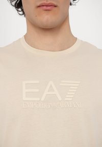 Krémové bavlnené tričko s vyvýšeným logom v strede: "EA7 EMPORIO ARMANI" v hladkom, textúrovanom písmene.