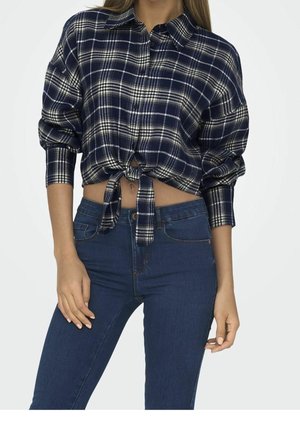 Vrouw met een marineblauw geruit overhemd met lange mouwen, geknoopt in de taille, en een high-rise blauwe jeans, staand tegen een lichte effen achtergrond.