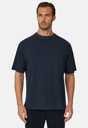Homme aux cheveux bruns courts et à la barbe, portant un t-shirt bleu marine uni à manches courtes et col rond, debout sur un fond blanc.