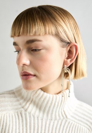 Metalløreringer med unikt buede design, som har flere kaskader af strenge og små metalperler. Modellen har lyst blont hår og bærer en hvid ribbet sweater.