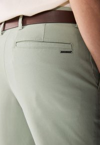 Un pantalon vert clair présente un design épuré avec une texture subtile, une poche arrière et une étiquette noire avec "Pier One" pour le marquage.