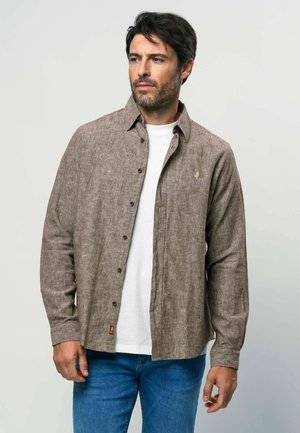 Muž s tmavými vlasy v hnědé košili na knoflíky přes bílé tričko a v modrých džínách, stojící před jednoduchým světlým pozadím.