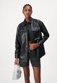 Giacca camicia nera in ecopelle con bottoni a pressione, due tasche frontali, dettagli in cucitura bianca, abbinata a pantaloni corti in denim neri e una borsa chiara.