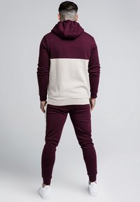 Ensemble de hoodie et de joggers en blocs de couleurs bordeaux et beige, fabriqué en tissu doux. Comprend une capuche, des poignets côtelés et une coupe fuselée aux chevilles.