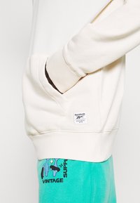 Krämfärgad sweatshirt med kenguruficka. Ribbad mudd vid ärmar och nederkant. Har en Reebok-logotyp och är ihop med tealfärgade sweatpants med grafiska tryck.