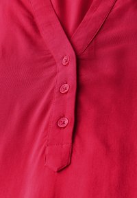 Rote Bluse mit V-Ausschnitt, ausgestattet mit fünf pinkfarbenen Knöpfen und leichten Falten auf der linken Seite, aus einem glatten, leichten Stoff gefertigt.