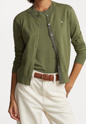 Personne portant un cardigan vert olive boutonné sur une chemise assortie, un pantalon blanc et une ceinture tressée marron avec une boucle dorée, main reposant dans la poche.