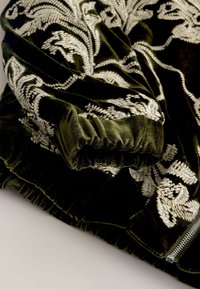 Velvet green jacket featuring intricate white embroidery, fitted cuffs, and a zip closure, showcasing rich texture and detailed patterns.
