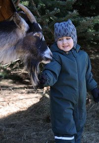 Niño con un mono impermeable verde oscuro con cremallera y franjas reflectantes interactúa con una cabra marrón y blanca en un entorno natural.