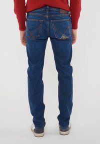 Uomo che indossa jeans blu con cuciture arancioni distintive e un maglione rosso, in piedi con le mani rilassate ai lati.