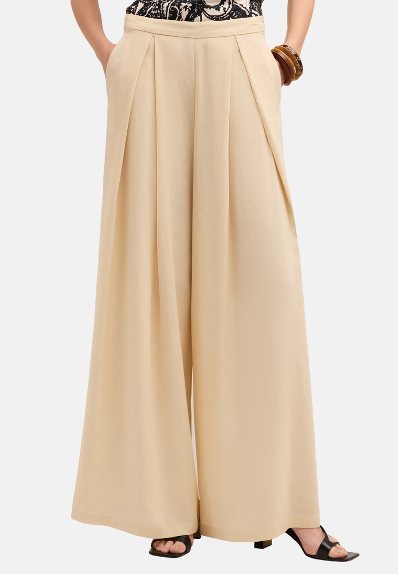 Pantaloni beige a gamba larga con design plissettato nella parte frontale, tasche laterali e una texture liscia, abbinati a sandali neri con punta aperta.