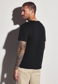 Muž s krátkými vlasy a kompletními tetováními na pažích, oblečený v černém tričku a béžových kalhotách, dívající se do strany na bílém pozadí.