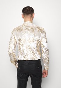 Camisa de manga larga en blanco con patrón floral dorado, confeccionada con tela suave. Se lleva con pantalones negros texturizados y detalle visible en la parte trasera.