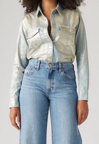 Camicia a maniche lunghe di un blu metallico con tasche anteriori e chiusura a bottoni, abbinata a jeans chiari a vita alta.