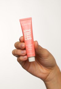 Rosa Quetschtube mit weißem Text, beschriftet mit "FREE STYLE Multi-Tasking Colour", ausgestattet mit einem weißen Deckel, in einer Hand vor einem hellen Hintergrund gehalten.