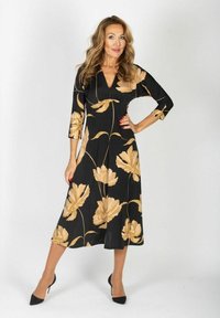 Mustat kukalliset mekko kultaisilla yksityiskohdilla, V-pääntie, kolme neljännestä hihaa, virtaava siluetti, puolipitkä, yhdistetty mustiin korkokenkiin.