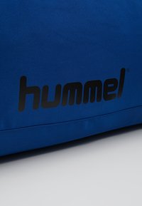 Bolsa azul hecha de un tejido duradero, con un gran logotipo negro de "hummel". Textura suave con un detalle de costura.
