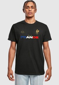 Mustan puuvillat-paidan etupuolella lukee "Vive la FRANCE" sinisissä, valkoisissa ja punaisissa kirjaimissa, ja rinnassa on kultainen kukkokuvio ja logo.