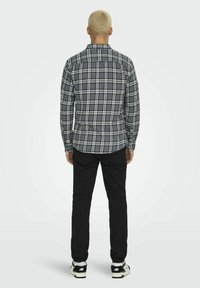 Grau kariertes Hemd mit langen Ärmeln und weißen Akzenten. Kombiniert mit schwarzen Hosen und Sneakers, von hinten betrachtet.