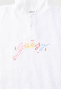 Λευκό τοπ με υφή υφάσματος, πολύχρωμο κεντημένο λογότυπο "guess" και κοντή λωρίδα με κουμπιά κάτω από τον στρογγυλό λαιμόκοψη.