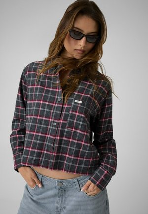 Femme portant une chemise à carreaux foncée avec des lignes roses et blanches, un jean bleu clair et des lunettes de soleil noires, debout les mains dans les poches.