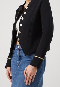 Schwarze cropped Jacke mit goldenen Knöpfen, strukturiertem Stoff und goldenen Akzenten an den Ärmeln; getragen über einem weißen Oberteil und einem blauen Jeansrock.