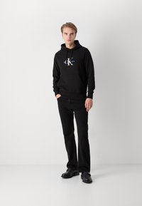 Felpa nera con logo grafico, tasca a marsupio e cordini, abbinata a jeans neri e scarpe nere, in piedi contro uno sfondo neutro.