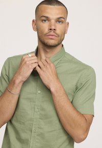 Homme aux cheveux courts et à la barbe ajustant le col d'une chemise à manches courtes vert clair, sur fond uni.